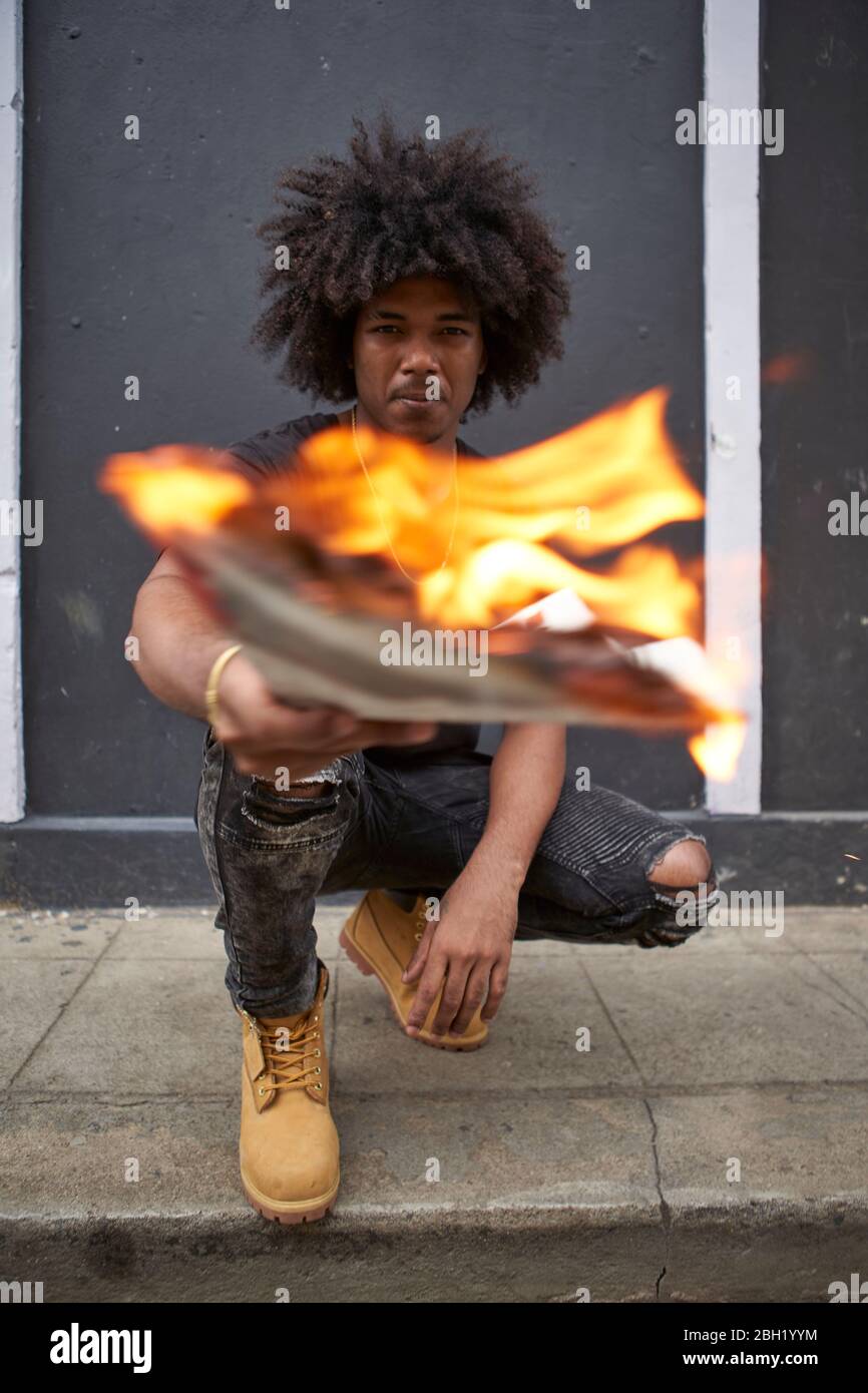 Ritratto di giovane uomo accovacciato sul marciapiede tenendo un giornale in fiamme Foto Stock