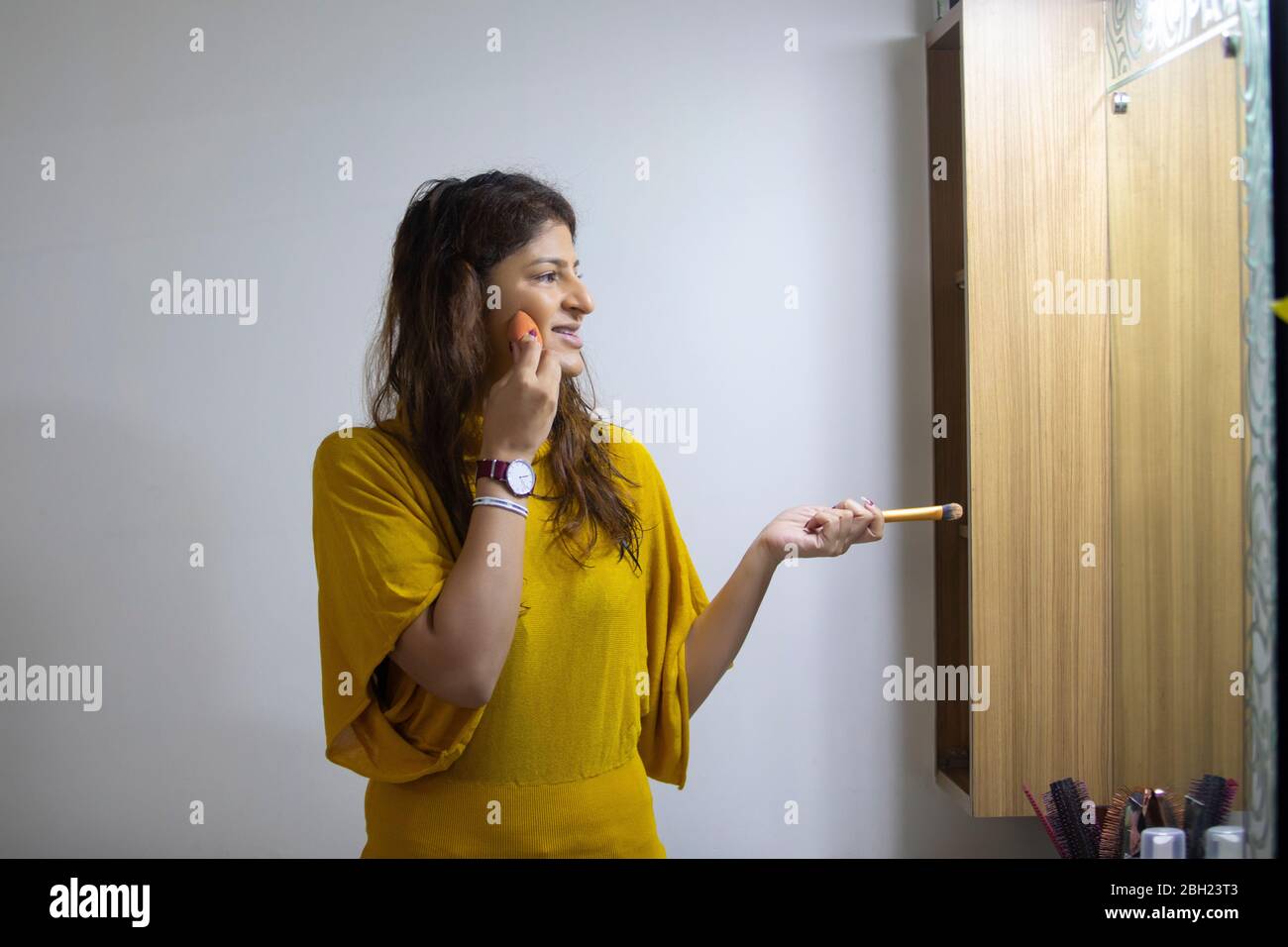 Donna che guarda nello specchio e che mescola il suo trucco mentre tiene una spazzola di trucco nella sua altra mano. Foto Stock