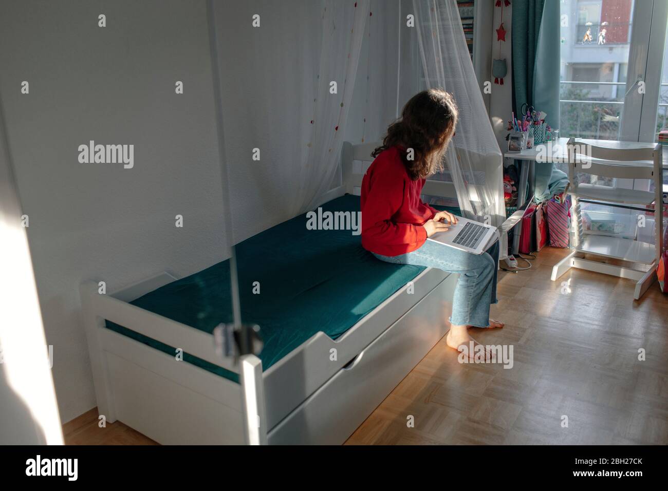 Ragazza con un computer portatile seduto sul letto nella camera dei bambini che guarda fuori dalla finestra Foto Stock