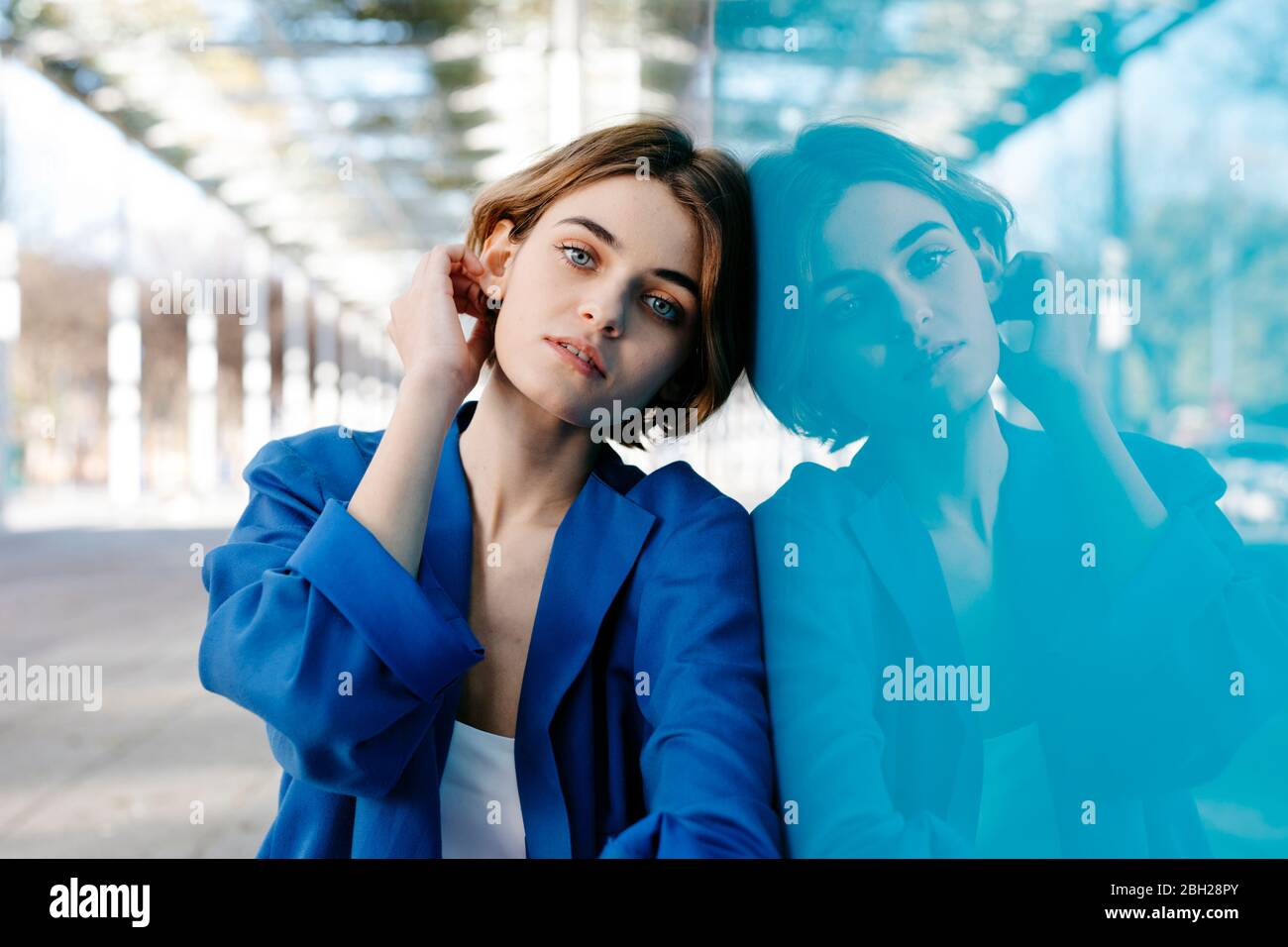 Giovane donna con aspetto urbano seduta sul pavimento e appoggiata su una colorata parete di vetro con il suo riflesso Foto Stock