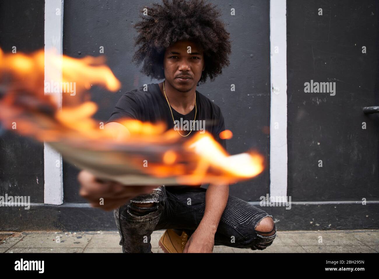 Ritratto di giovane uomo accovacciato sul marciapiede tenendo un giornale in fiamme Foto Stock