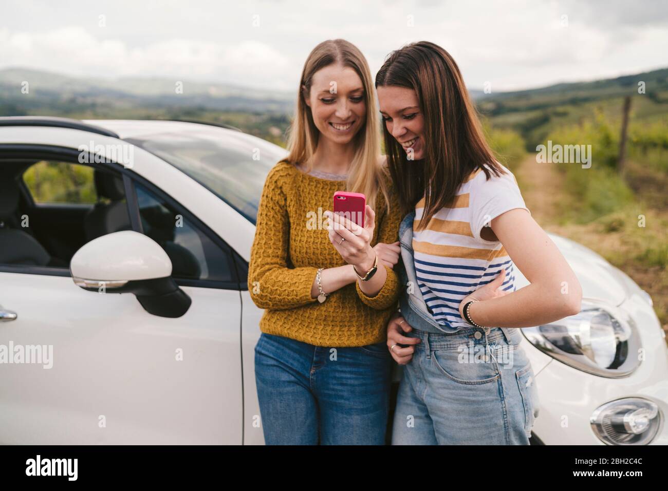 Due giovani donne felici in piedi accanto auto che condividono telefono cellulare Foto Stock