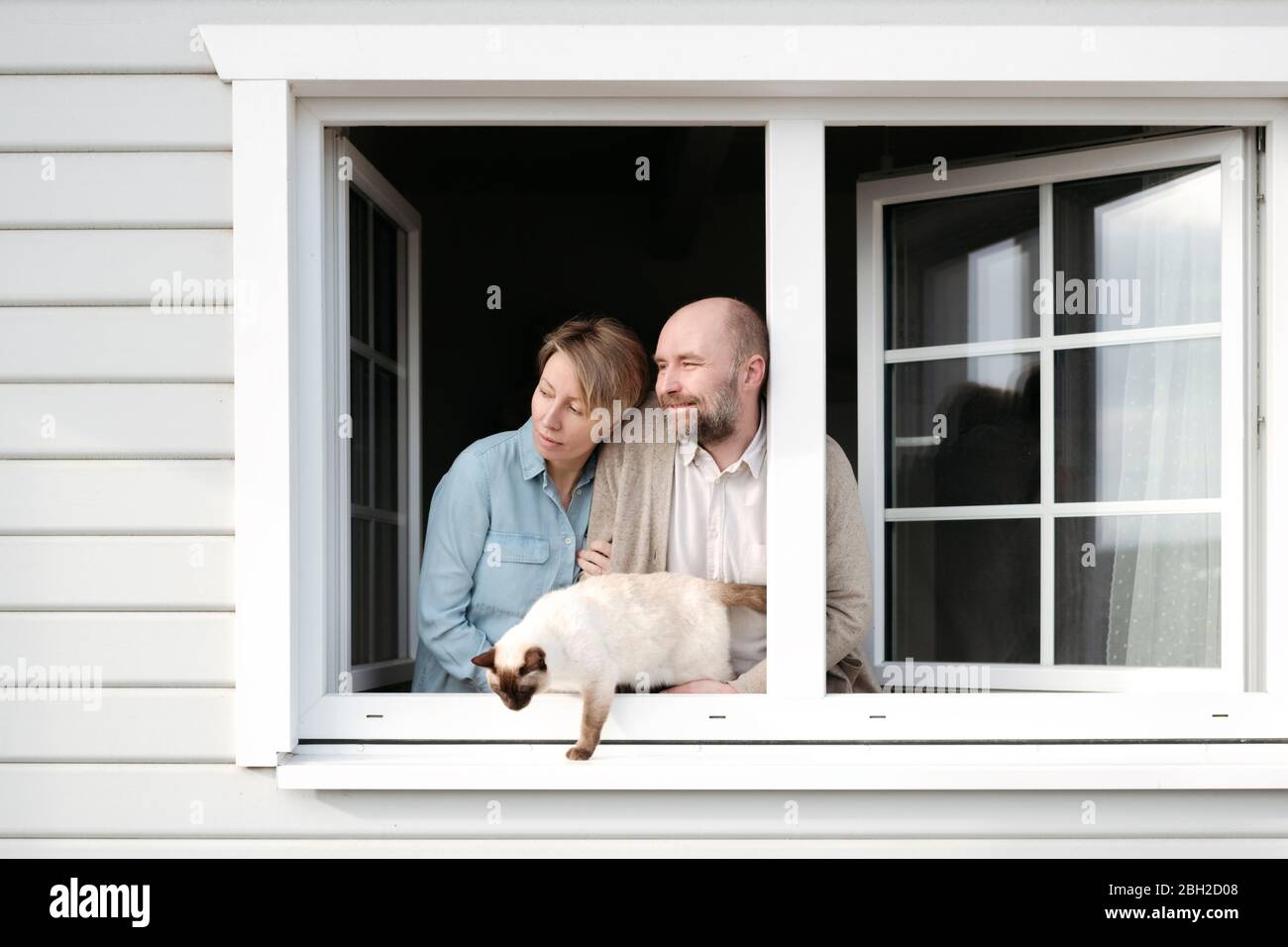 Coppia con gatto che si appoggia fuori dalla finestra della loro casa Foto Stock