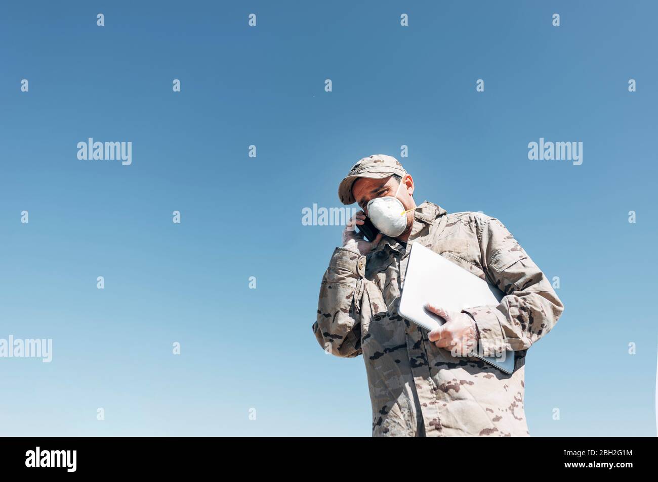 Soldato con maschera facciale in caso di emergenza, utilizzando smartphone, trasportando un computer portatile Foto Stock