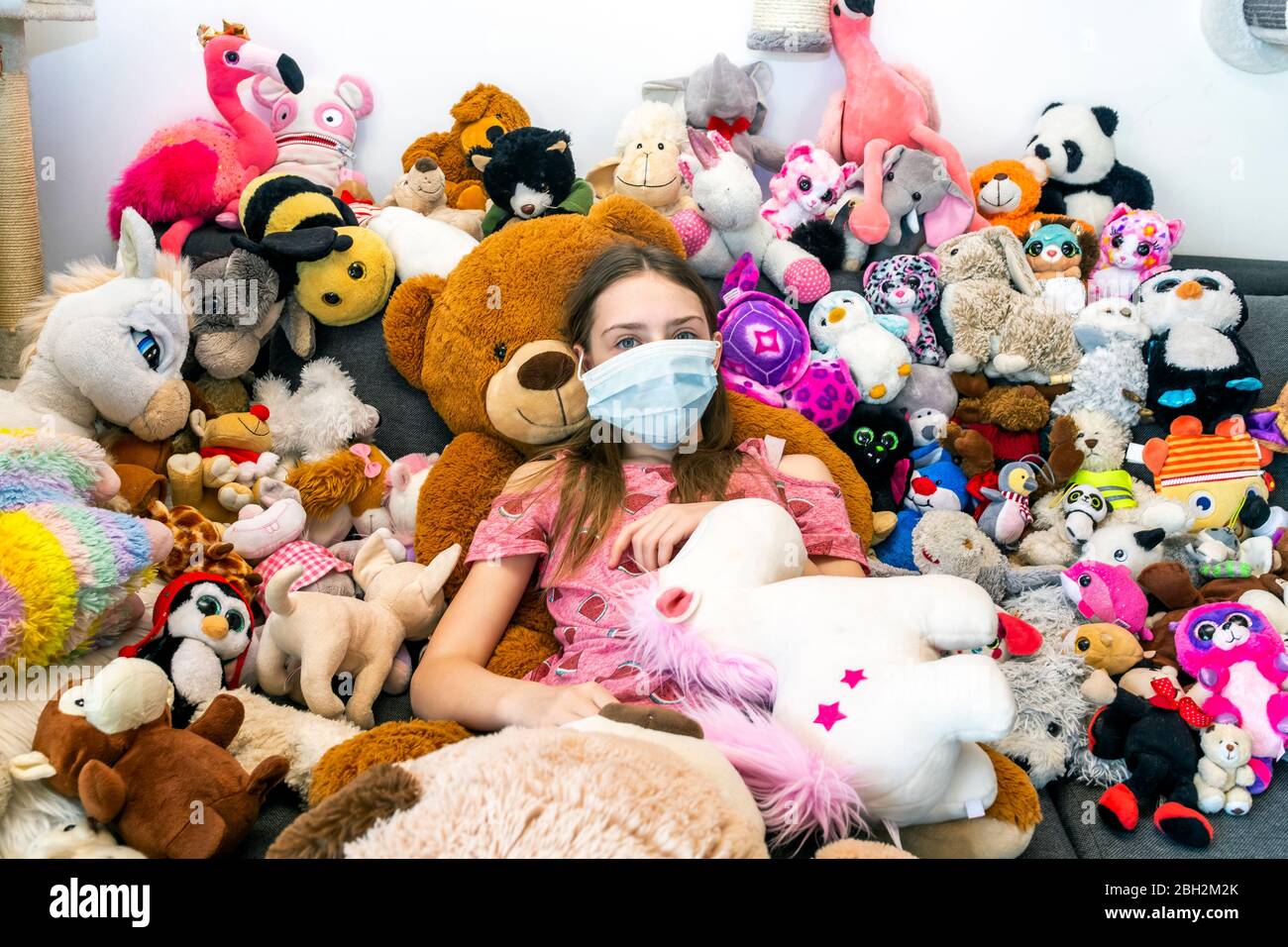 Ragazza con maschera facciale, seduta sul divano, coperta di giocattoli coccolati Foto Stock