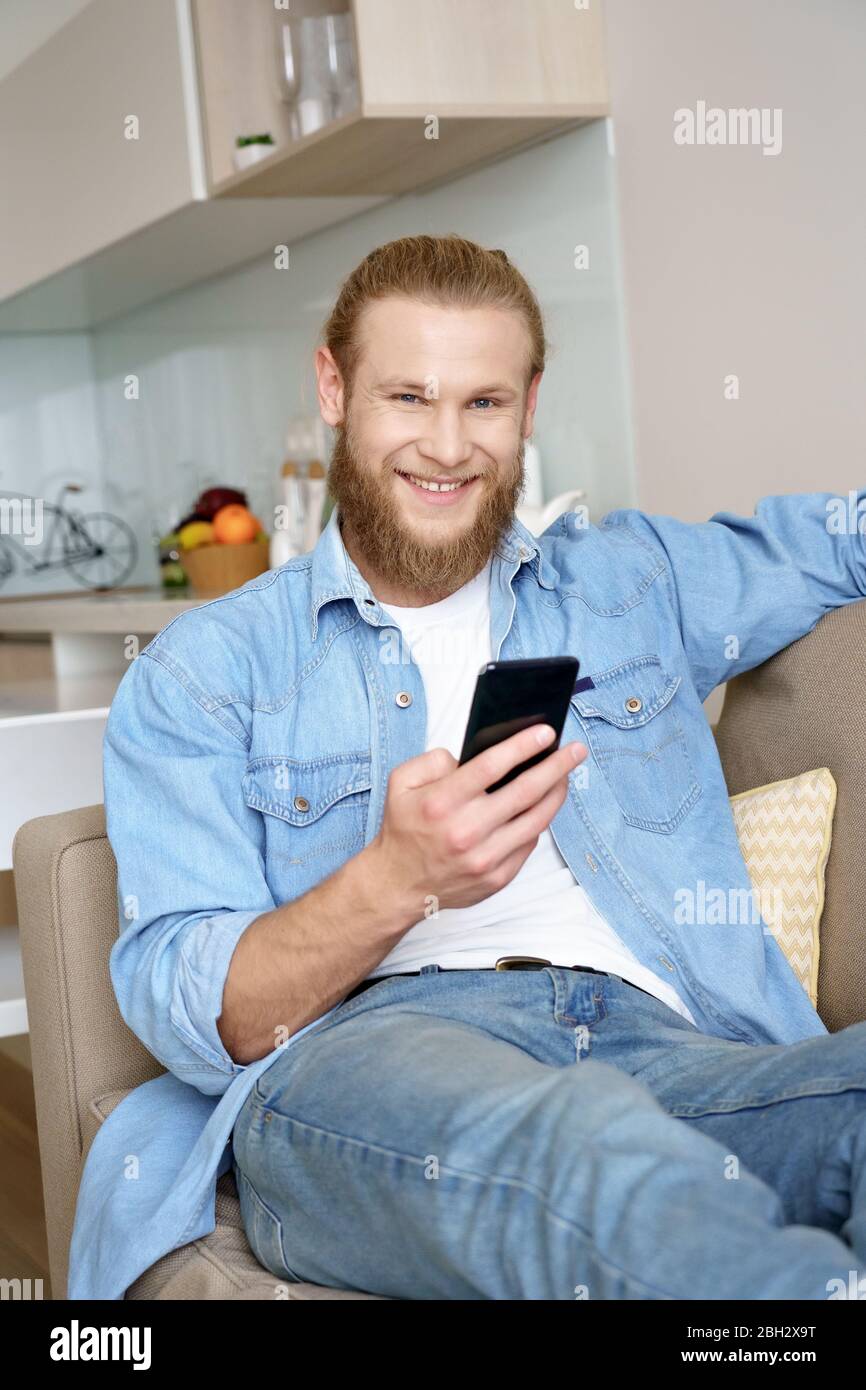 Buon giovane uomo che si rilassa sul divano utilizzando lo smartphone guardando la fotocamera a casa. Foto Stock
