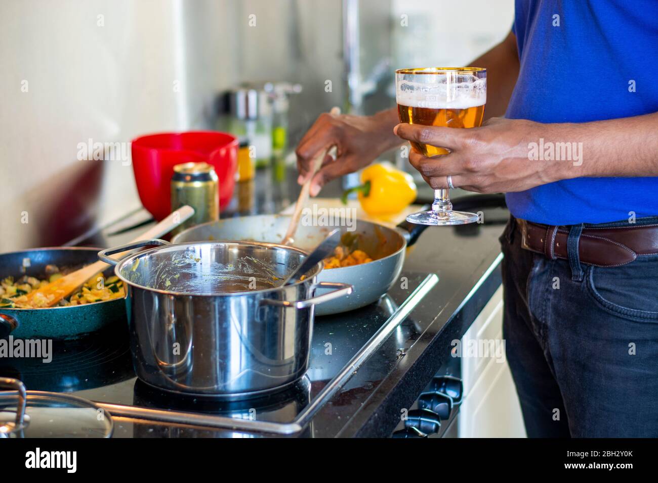 Uomo che cucina su una stufa elettrica in cucina mentre beve una birra. Preparazione del cibo. Bancone cucina caotico Foto Stock