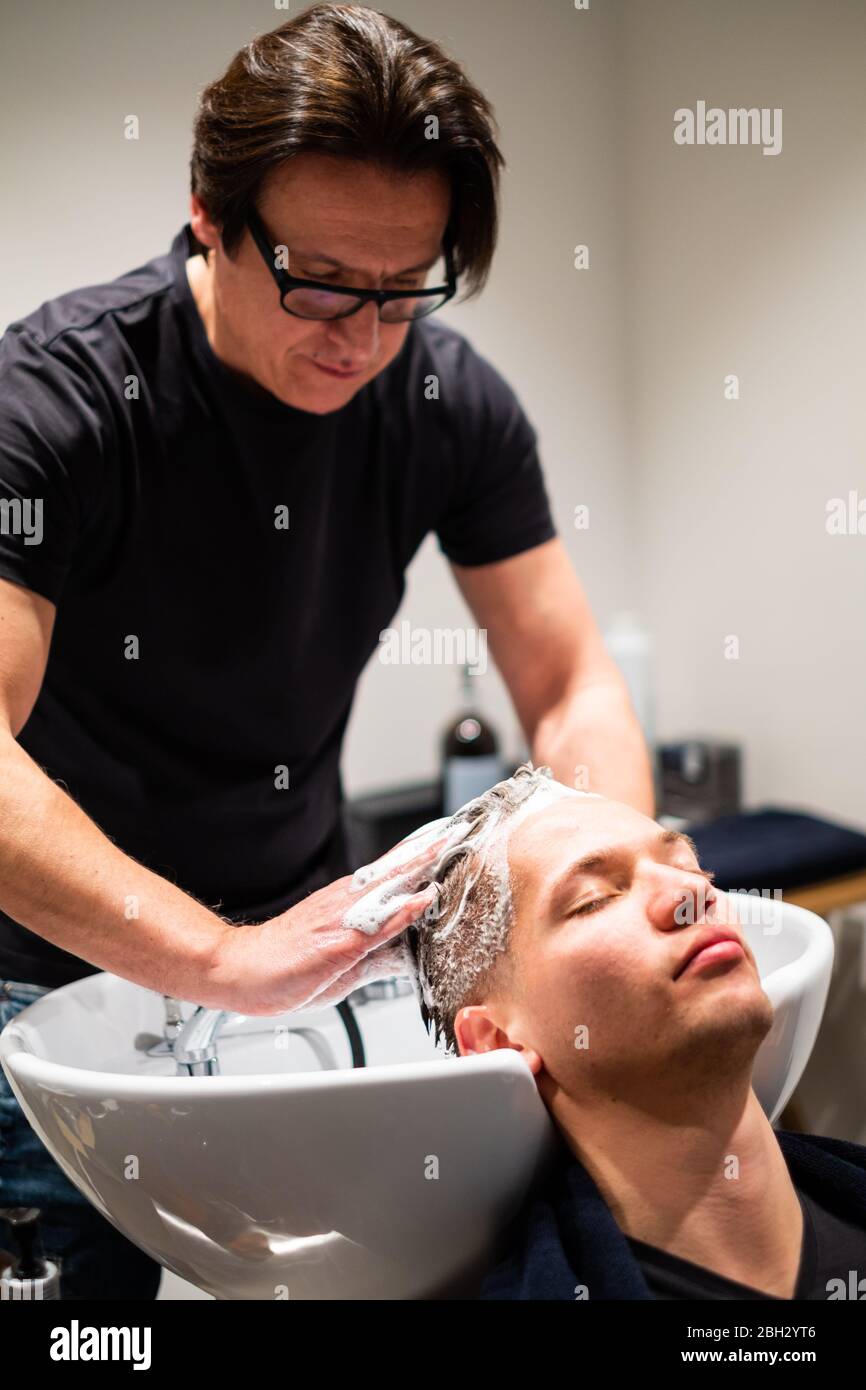 Barber lava i capelli del giovane con molta schiuma nel suo barbiere. Massaggia la testa sul lavandino. Foto Stock