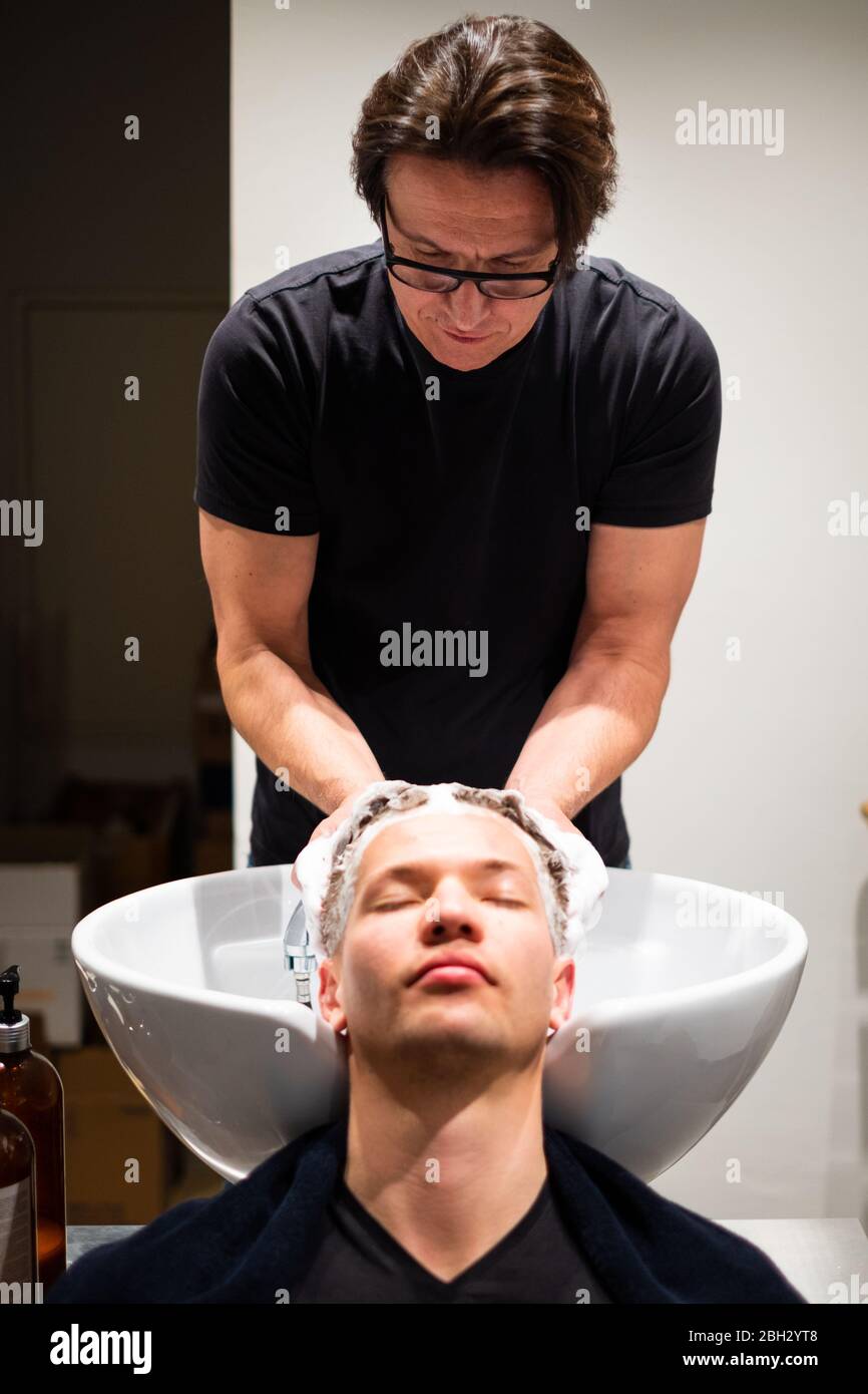 Barber lava i capelli del giovane con molta schiuma nel suo barbiere. Massaggia la testa sul lavandino. Foto Stock
