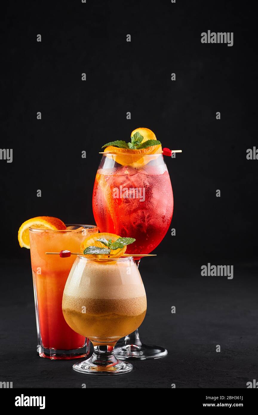 Tre cocktail alcolici, rossi, arancioni e gialli sullo sfondo nero. Varietà di bevande alcoliche decorate con fette di arancia. Spazio di copia Foto Stock