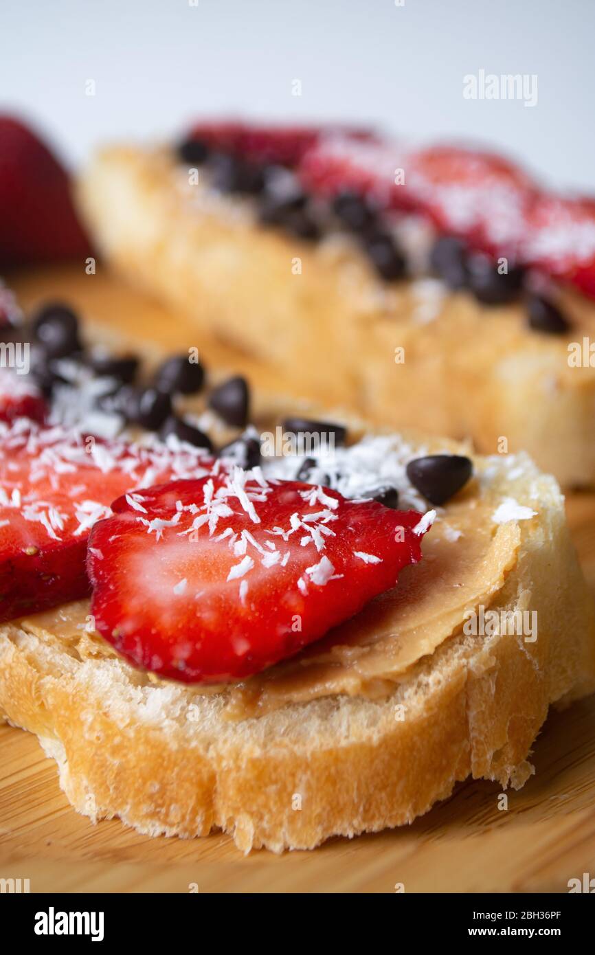Burro di arachidi spalmato con scaglie di cioccolato, fragole, scaglie di cocco e pane fatto in casa su un piatto di legno Foto Stock