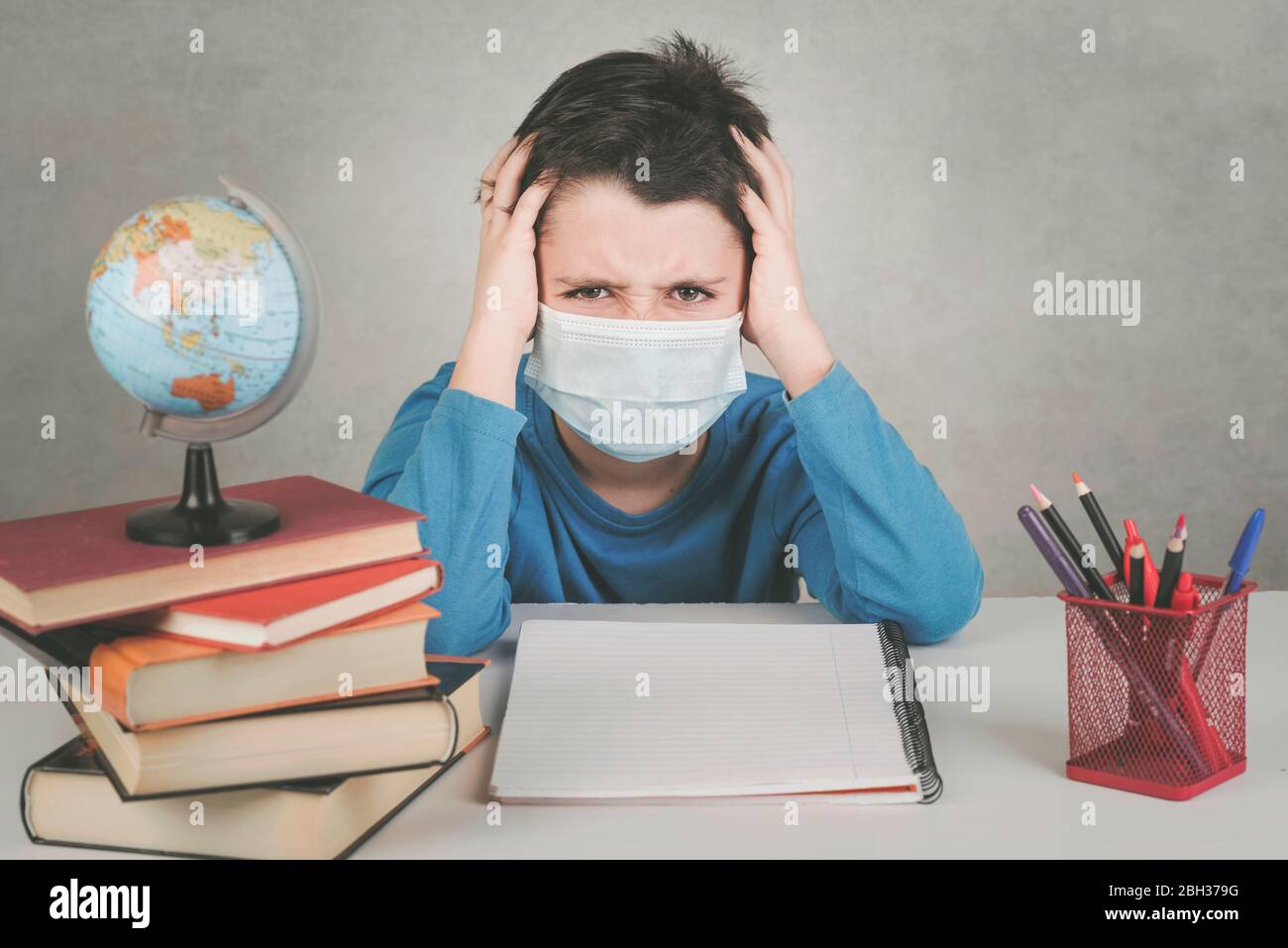 Bambino arrabbiato che indossa la maschera medica si è stuperso di fare i compiti nella quarantena su sfondo grigio Foto Stock