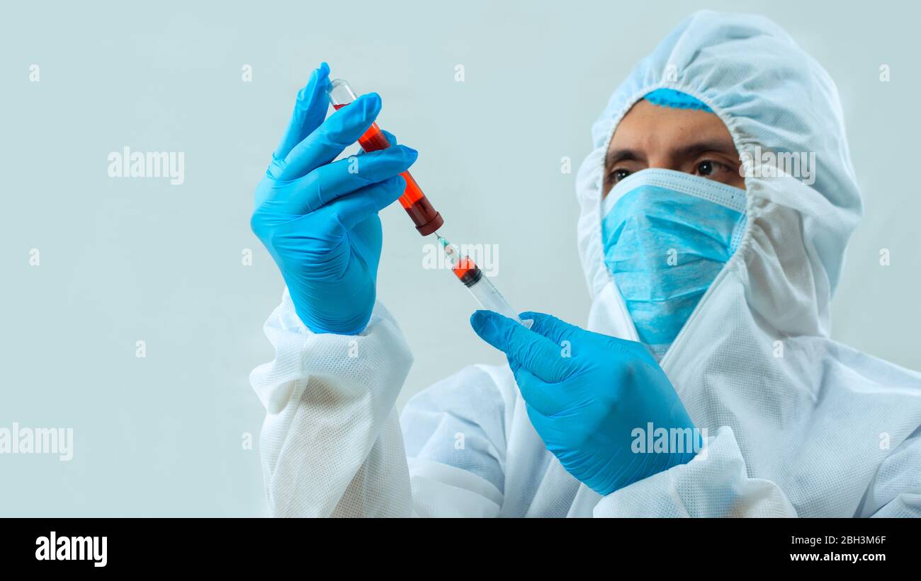Medico latino in tuta bioprotettiva, maschera facciale e guanti blu, che preleva un campione di sangue in una provetta con una siringa su sfondo bianco Foto Stock