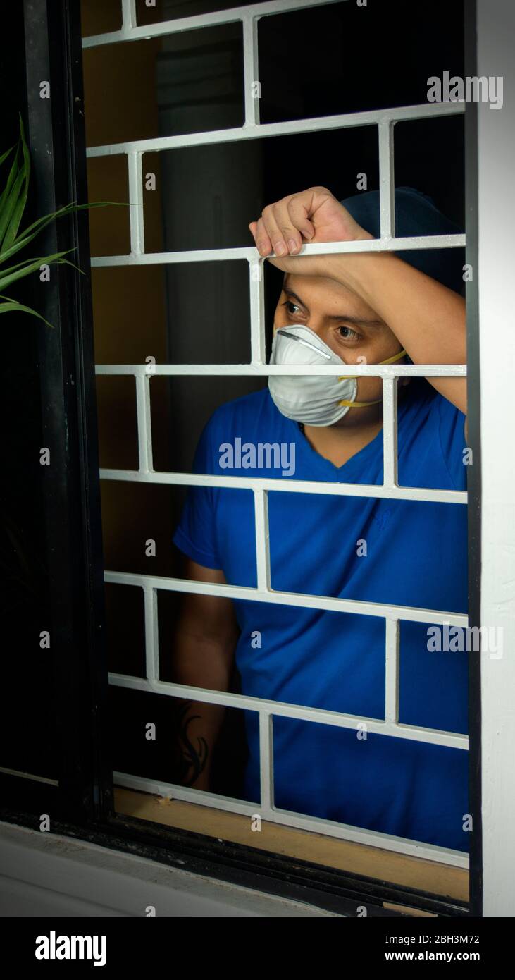 Latino uomo con maschera facciale bianca e camicia blu all'interno della sua casa guardando fuori attraverso la finestra, appoggiandosi contro le barre di sicurezza Foto Stock