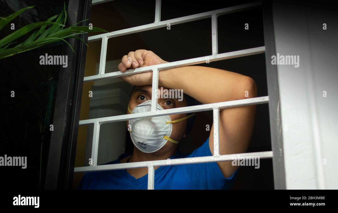 Latino uomo con maschera facciale bianca e camicia blu all'interno della sua casa guardando fuori attraverso la finestra, appoggiandosi contro le barre di sicurezza Foto Stock