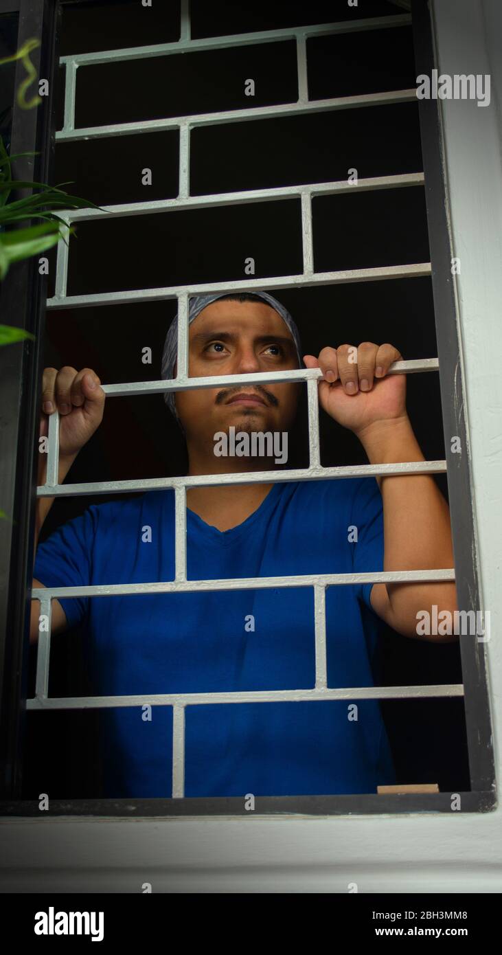 Vista ravvicinata dell'uomo latino con camicia blu chiusa a casa, che guarda fuori dalla finestra, appoggiandosi contro le barre di sicurezza Foto Stock
