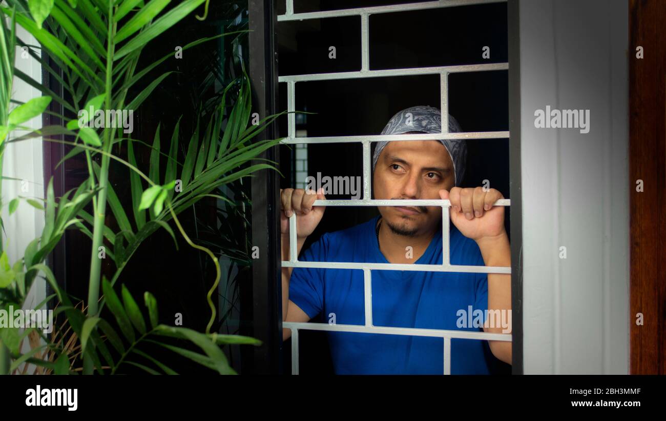 Vista ravvicinata dell'uomo latino con camicia blu chiusa a casa, che guarda fuori dalla finestra, appoggiandosi contro le barre di sicurezza Foto Stock