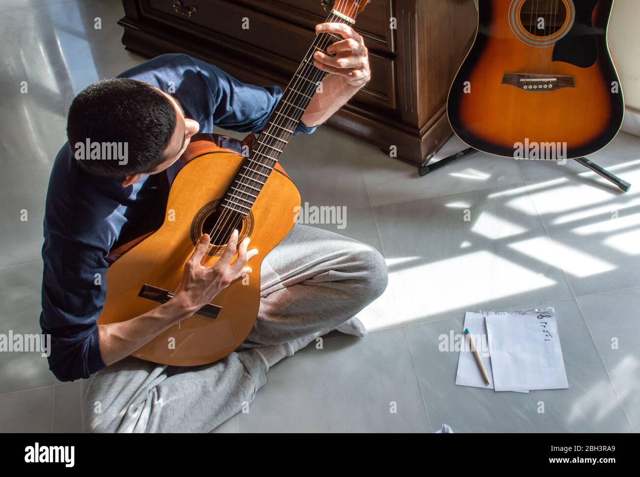 Uomo che suona la chitarra e compone musica a casa vicino a una finestra luminosa in una giornata di sole. Musicista informale seduto sul pavimento che suona la chitarra. Foto Stock