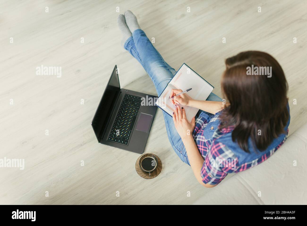 La ragazza seduta a casa sul pavimento con un computer portatile, una tazza di caffè e fare diario entries.Homeworking, freelance. Distanza sociale Concept.Quaranti Foto Stock