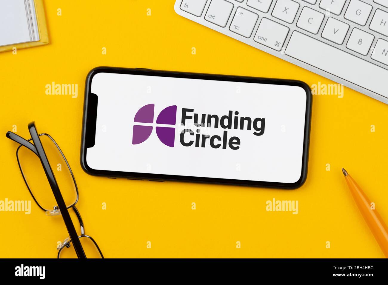 Uno smartphone con il logo Funding Circle si trova su uno sfondo giallo insieme a tastiera, occhiali, penna e libro (solo per uso editoriale). Foto Stock