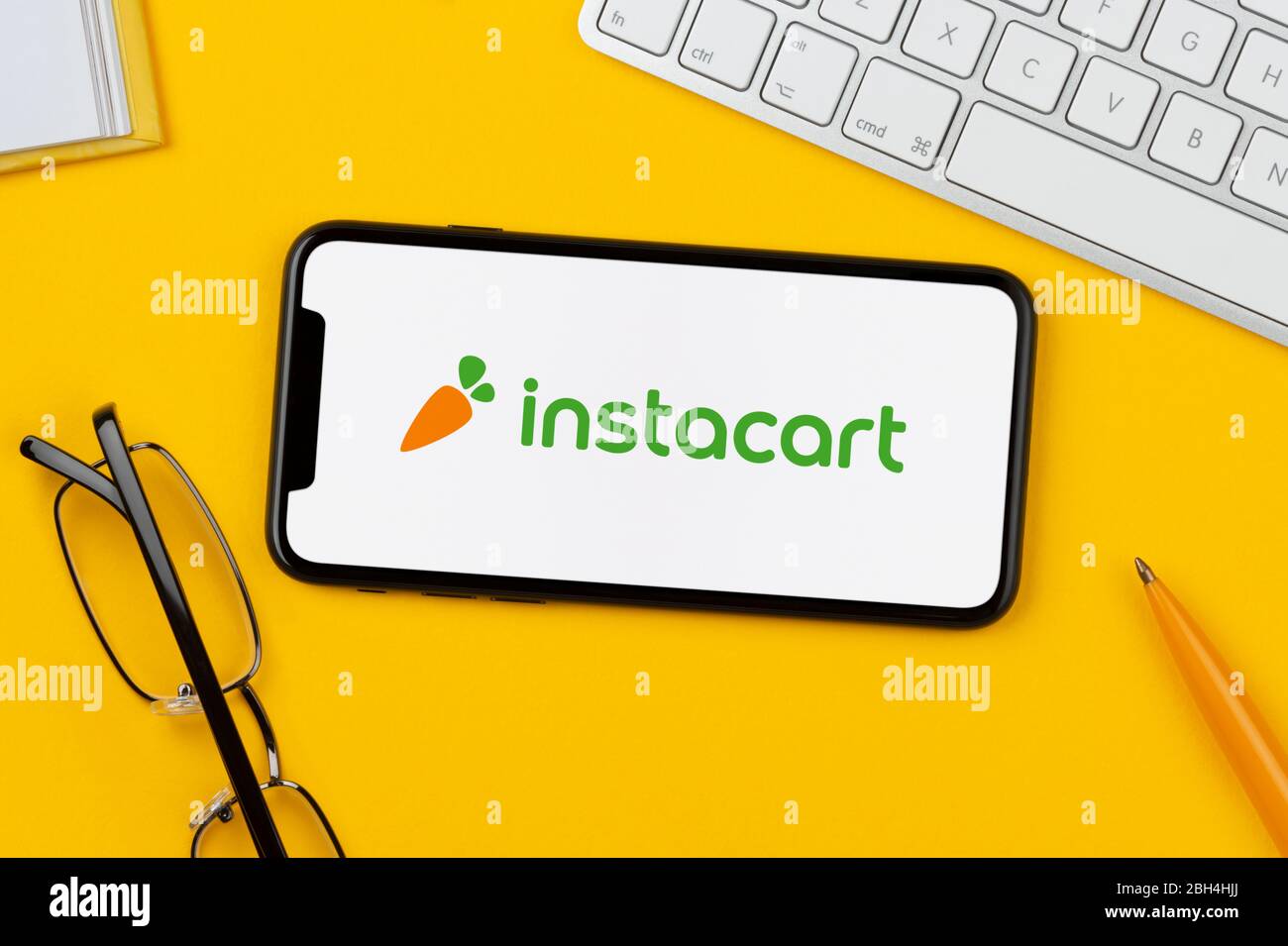 Uno smartphone con il logo Instacart è posizionato su uno sfondo giallo insieme a tastiera, occhiali, penna e libro (solo per uso editoriale). Foto Stock
