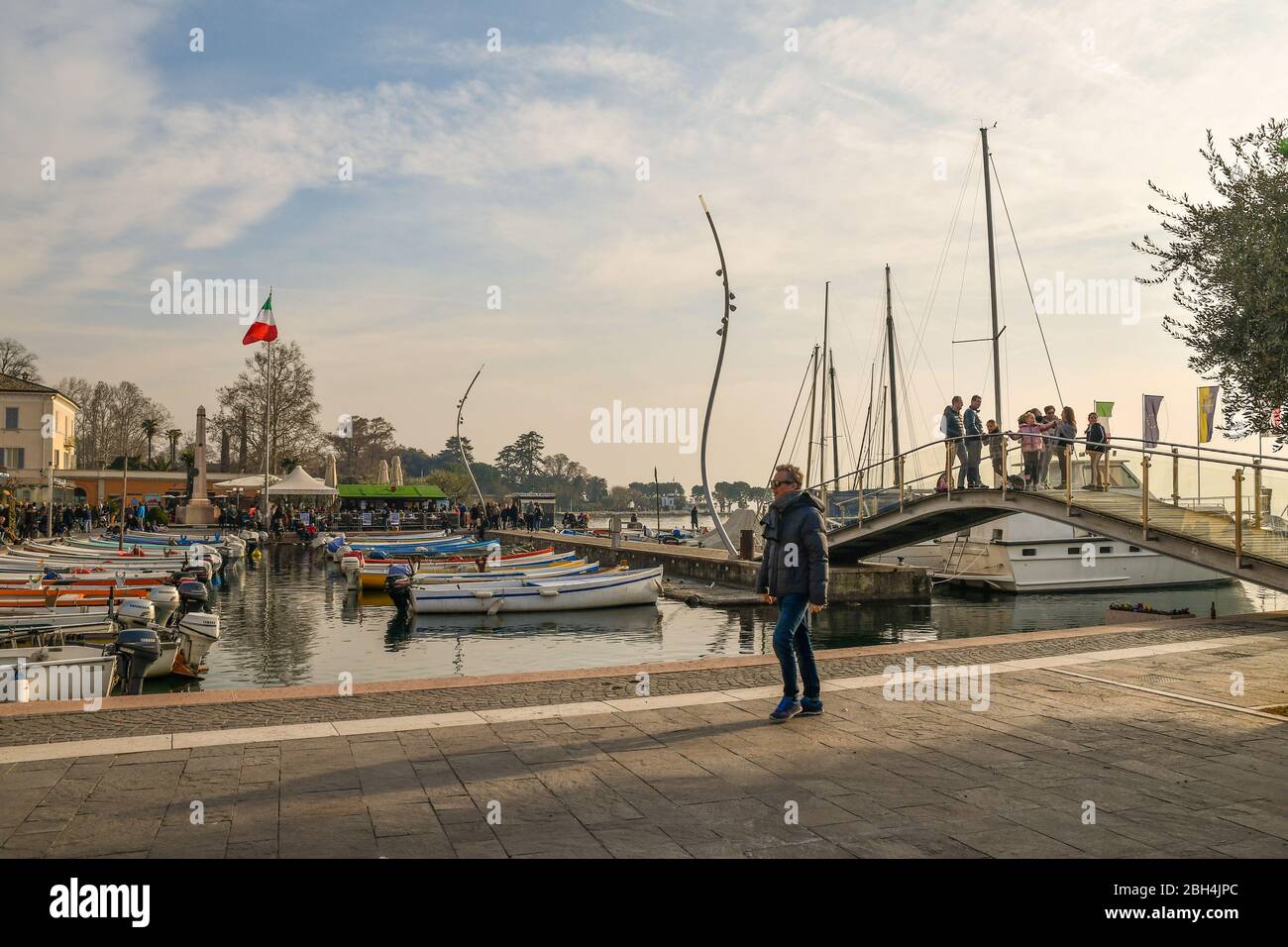 Porto del centro storico sulla riva del Lago di Garda con barche ormeggiate, un ponte pedonale e la gente ad un festival sullo sfondo, Bardolino, Italia Foto Stock