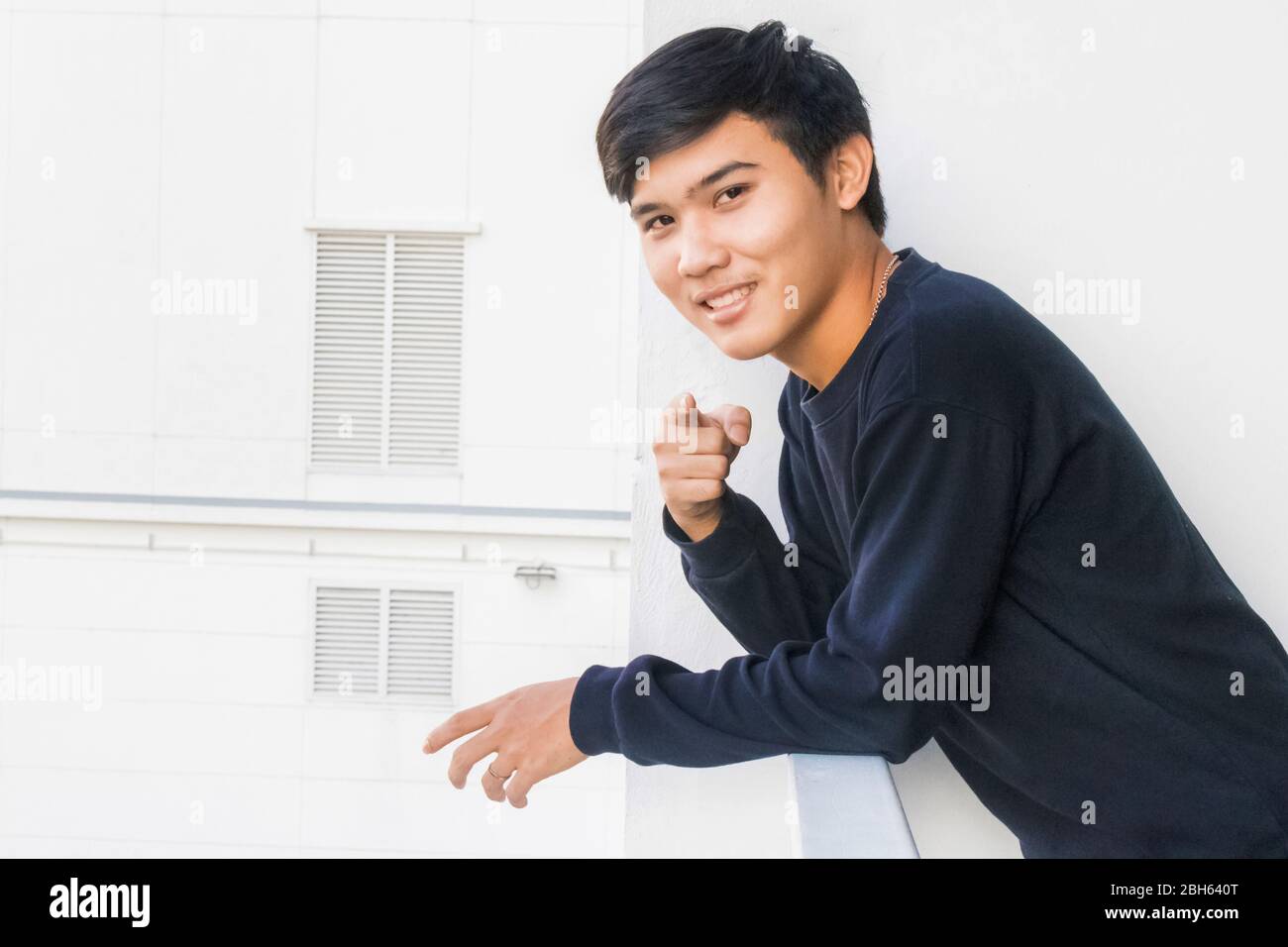 Un ragazzo tailandese in abiti neri in piedi nella terrazza bianca e punta alla macchina fotografica. Foto Stock
