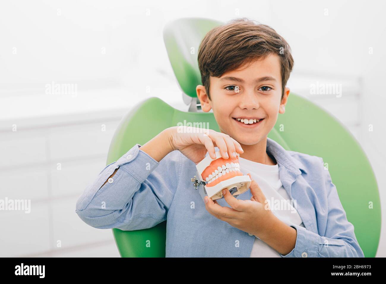 Ragazzo di razza mista che tiene la mascella dentale mentre siede in sedia dentale. È felice dopo il trattamento dei denti Foto Stock