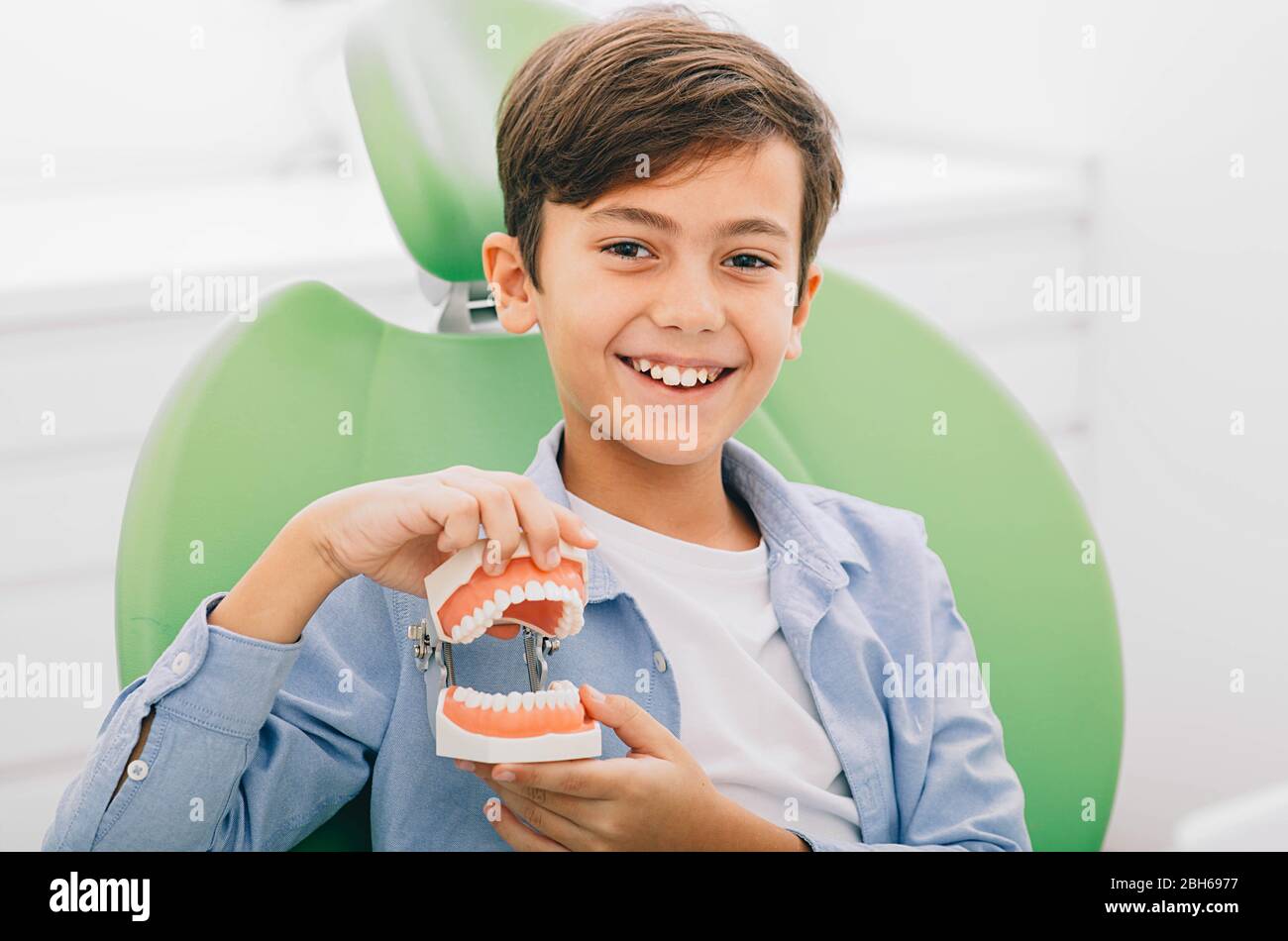 Ragazzo di razza mista che tiene la mascella dentale mentre siede in sedia dentale. È felice dopo il trattamento dei denti Foto Stock