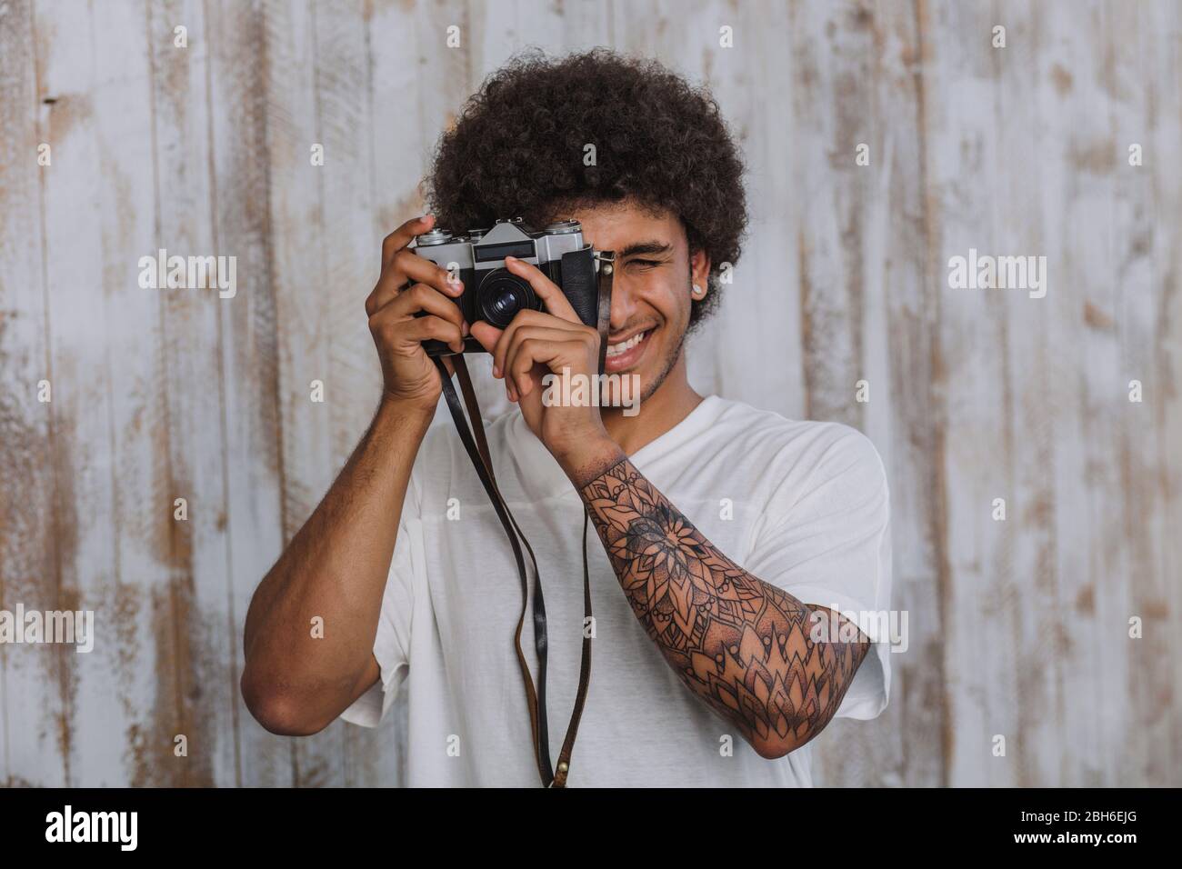 Attraente e uomo africano con capelli ricci, tenendo in mano una videocamera vicino al viso e fotografie, dietro di lui il vecchio sfondo di legno Foto Stock