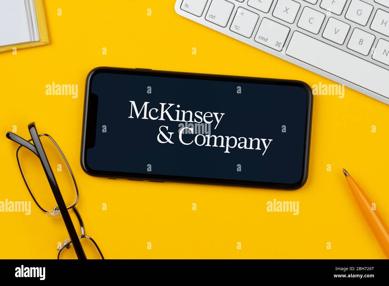 Uno smartphone con il logo McKinsey & Company si trova su uno sfondo giallo, insieme a tastiera, occhiali, penna e libro (solo per uso editoriale). Foto Stock
