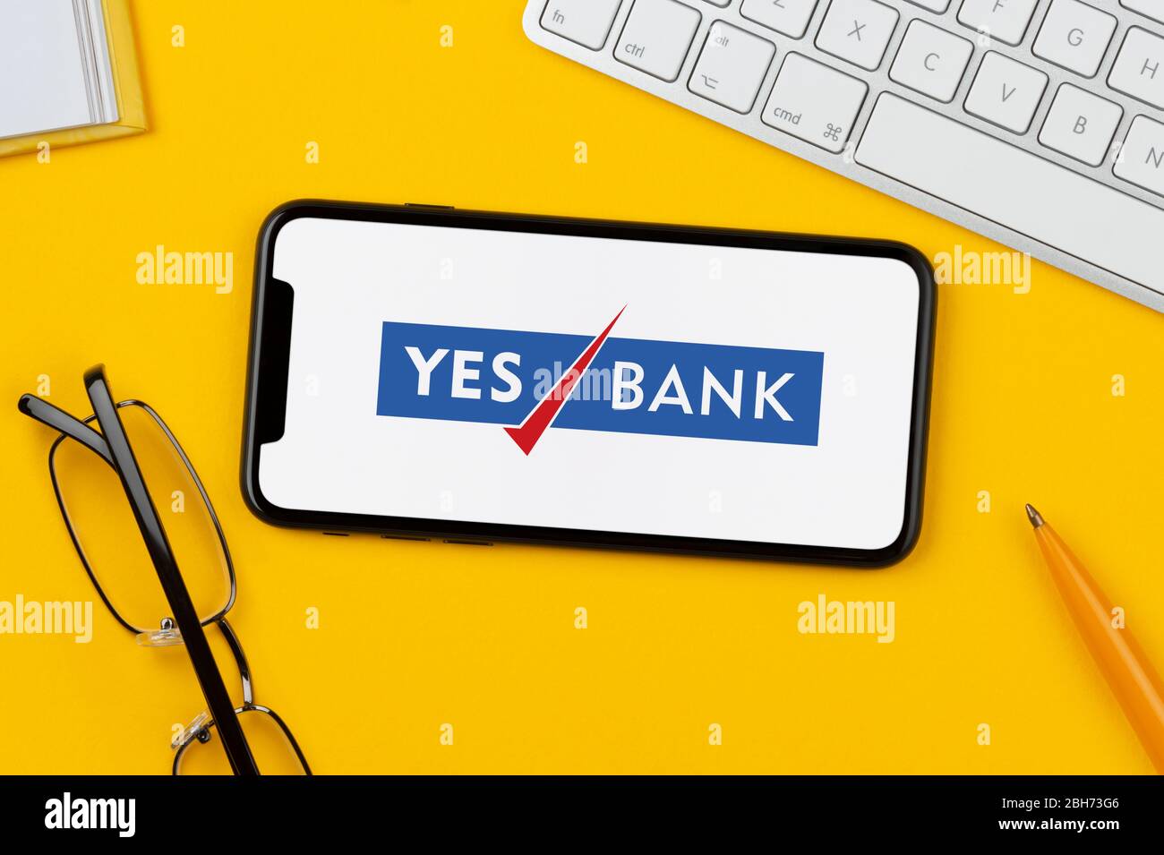 Uno smartphone con il logo Yes Bank è posizionato su uno sfondo giallo insieme a tastiera, occhiali, penna e libro (solo per uso editoriale). Foto Stock