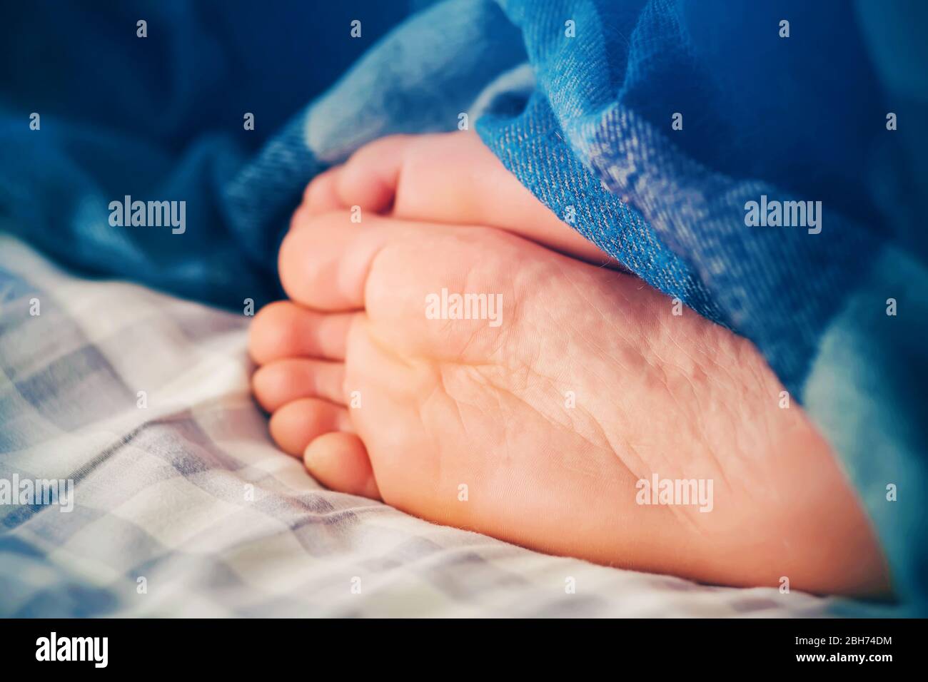 Le gambe di un uomo che dorme giacciono su un lenzuolo a scacchi sul letto, coperto da una coperta blu brillante e illuminato dalla luce al mattino. Foto Stock