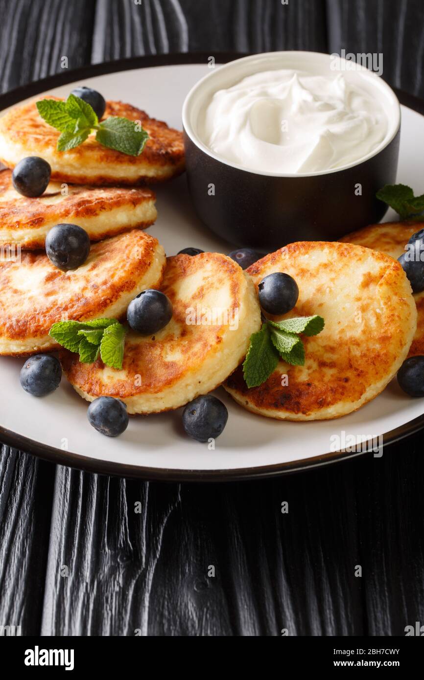 Colazione sana di sciroppi di formaggio casolare con mirtilli, menta e crema acida primo piano in un piatto sul tavolo. Verticale Foto Stock