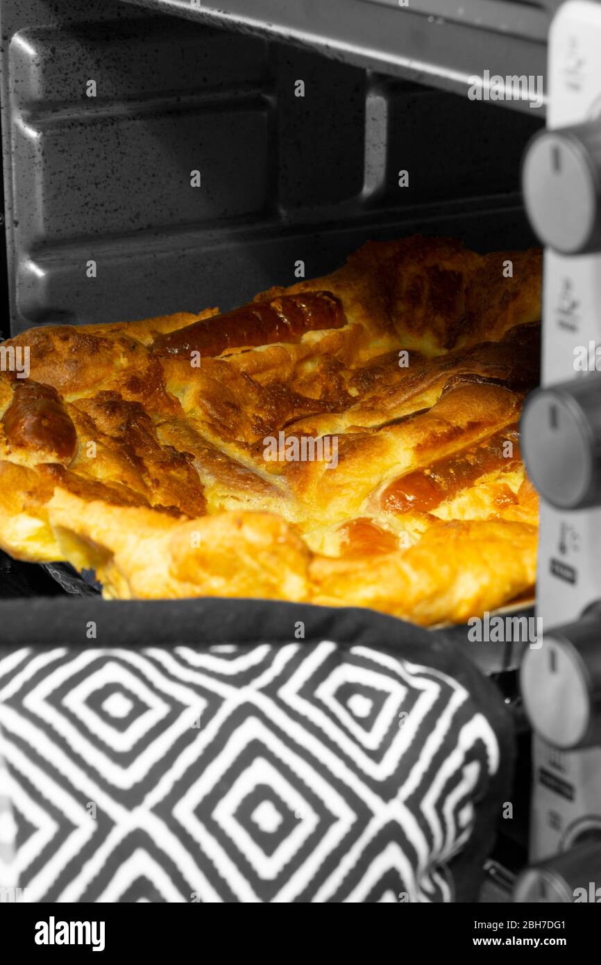 Toad nel buco, budino Yorkshire con salsiccia di maiale, che viene preso da un forno da una persona che indossa guanti da forno. Immagine a colori selettiva Foto Stock