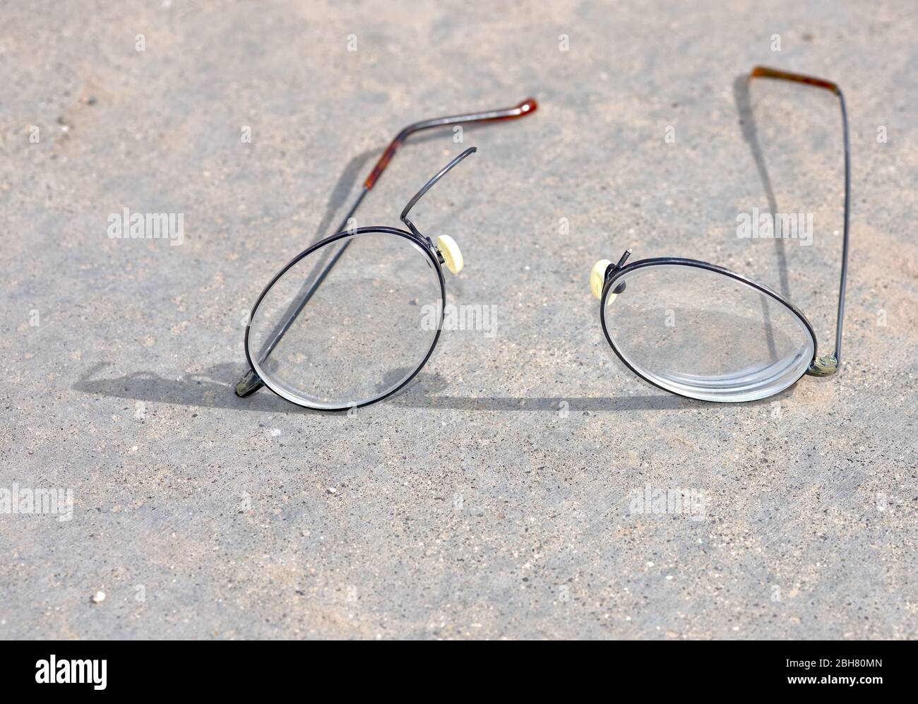 Un paio di occhiali con bordo in filo di titanio che si sono rotti e separati sulla sezione del ponte nasale mentre si posano su una superficie di cemento. Foto Stock