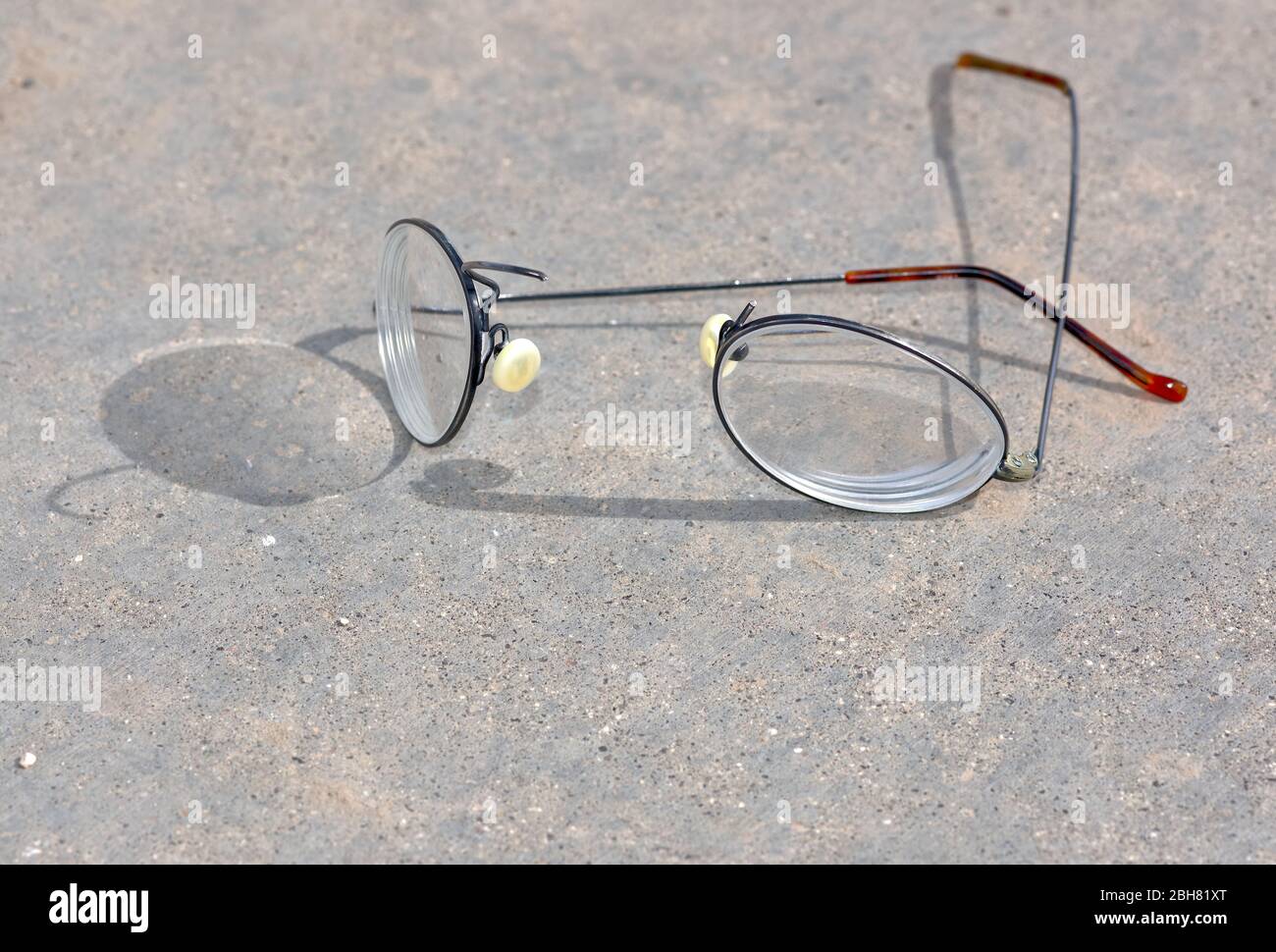 Un paio di occhiali con bordo in filo di titanio che si sono rotti e separati sulla sezione del ponte nasale mentre si posano su una superficie di cemento. Foto Stock