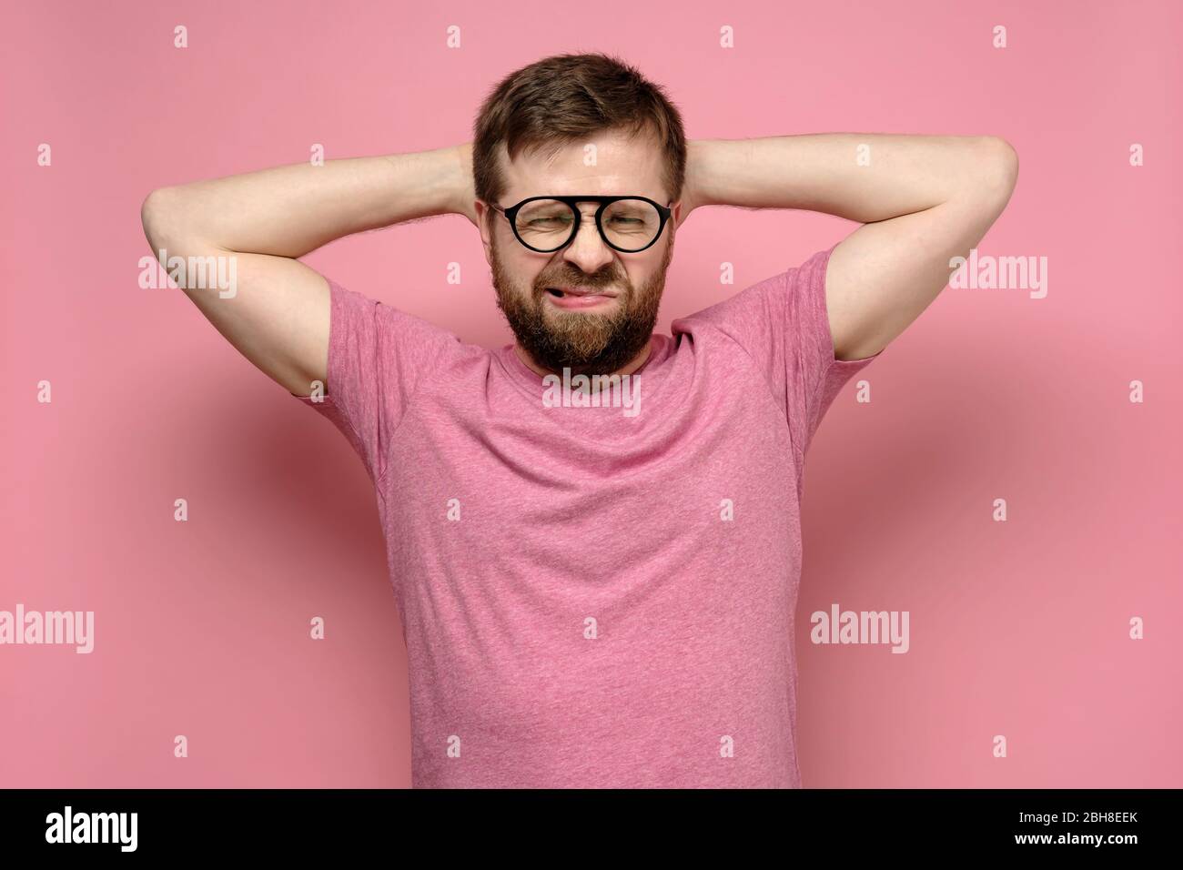 L'uomo bearded con gli occhiali non è nello spirito, lui grida nell'esasperazione, mettendo le mani dietro la testa, su uno sfondo rosa. Foto Stock