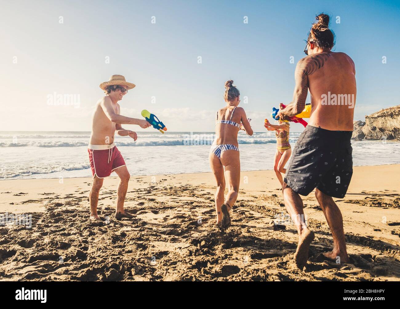 Gruppo di persone giovani caucasici uomini e donne giocano con pistola ad acqua in spiaggia durante le vacanze con gli amici insieme all'aperto in una giornata di sole vacanze - attiva giovane alternativa millenario Foto Stock