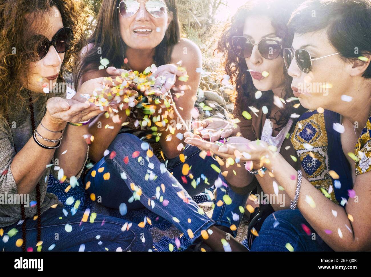 Gruppo di belle donne di mezza età giovani che giocano e soffiano un sacco di confetti colorati in aria - festeggiare e divertirsi insieme con l'amicizia - partito all'aperto concetto con retroilluminazione del sole Foto Stock