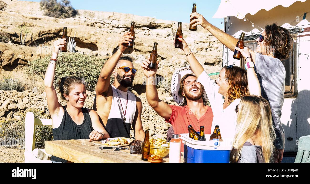 Divertimento e concetto di amicizia con un gruppo di giovani uomini e donne insieme tostando con birre e cibo seduti su un tavolo di legno rurale con la natura in background Foto Stock