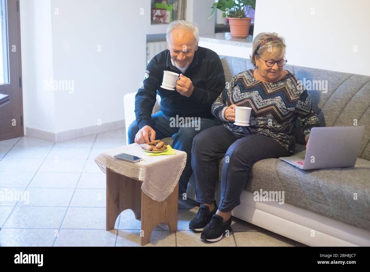 Pensionati anziani matura godere l'inverno in un pomeriggio guardando il computer portatile e di bere il tè seduto sul divano di casa. Tecnologia e concetto di piacere Foto Stock