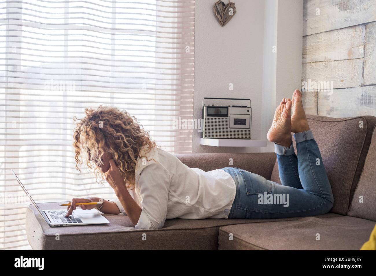 Donna curly a casa si stendono e utilizzano computer portatile sul divano in posizione inversa - lavoro al coperto in ufficio alternativo - casalinga gestione e fare affari - libero stile di vita delle persone - moderna tecnologia internet Foto Stock