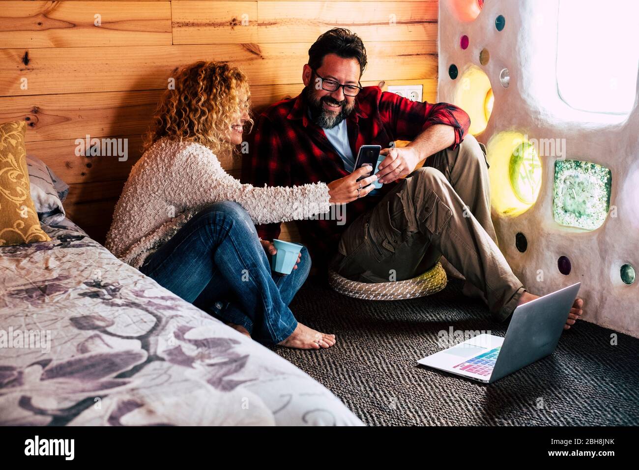 Coppia di adulti che gode di tecnologia a casa o in hotel che viaggia guardando uno schermo di telefono cellulare e utilizzando un computer portatile internet connesso al piano - concetto persone alternative Foto Stock