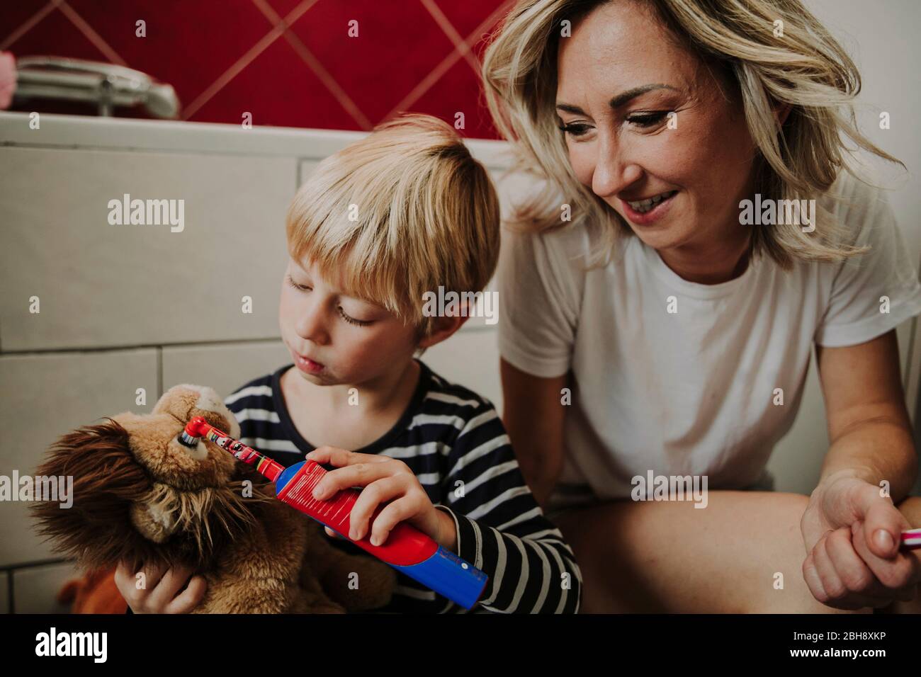kleiner Junge mit Stofftier und Zahnbürste, Mutter sieht ihm zu Foto Stock