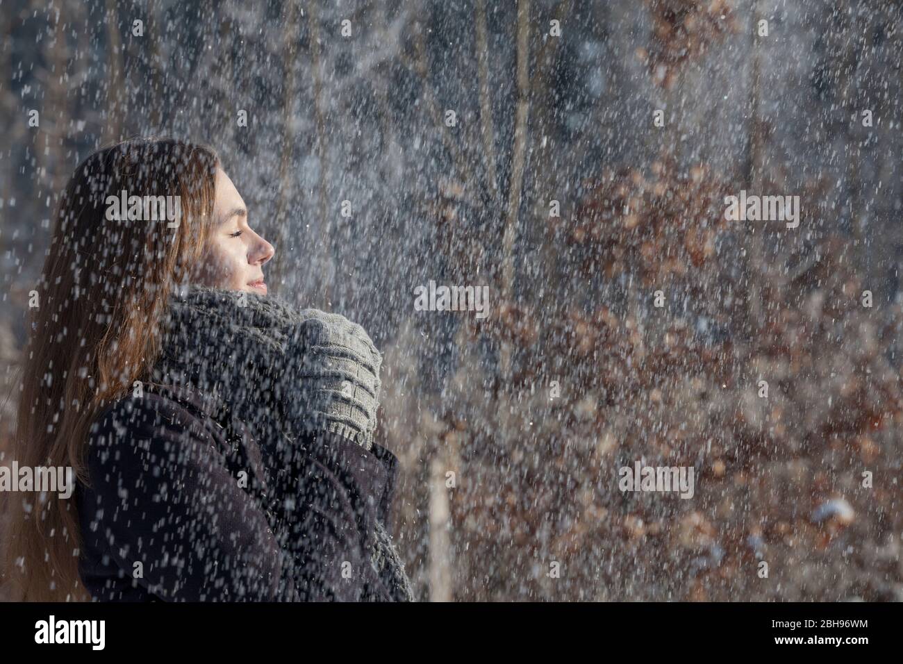Giovane donna gode con occhi chiusi il sole sul viso e la neve che cade, capelli lunghi marroni, abiti invernali, sciarpa e guanti Foto Stock