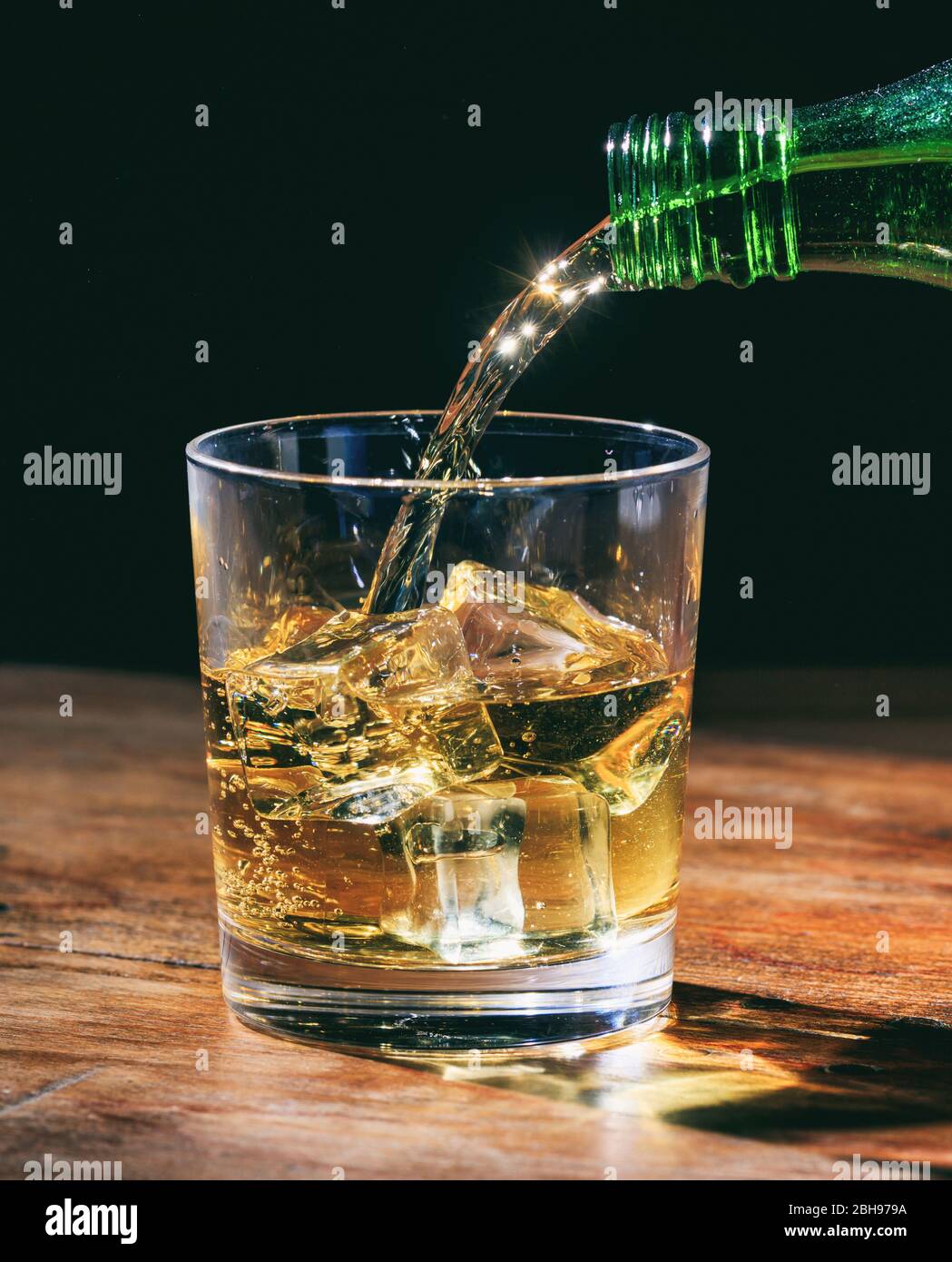 Bottiglia verde che versa whisky sulle rocce, in cristallo su tavolo di legno. Ritratto verticale della bevanda alcolica con cubetti di ghiaccio su sfondo scuro. Foto Stock