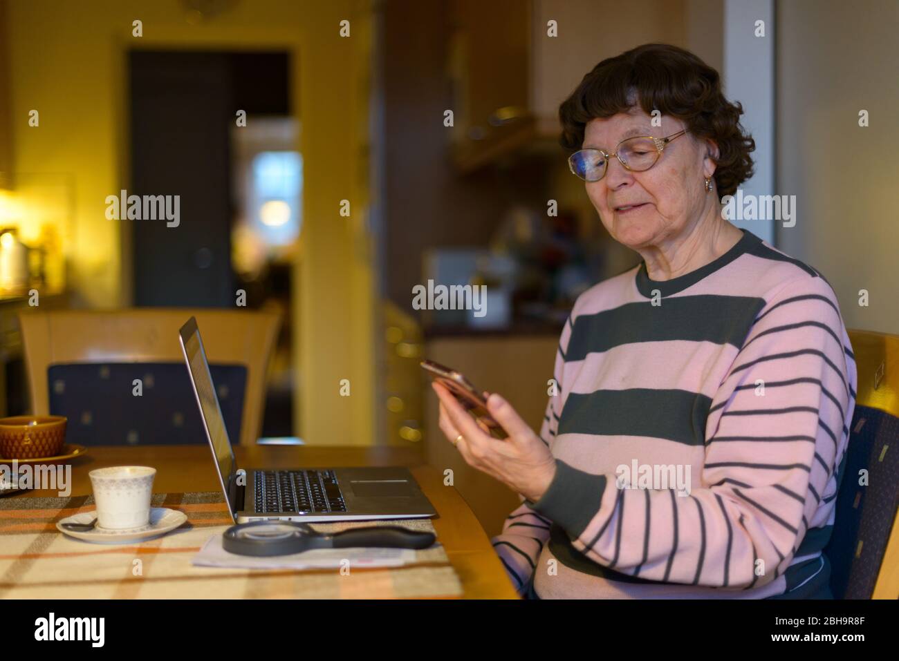 Felice donna senior sorridente mentre tiene il telefono cellulare con il computer portatile su un tavolo di legno nella sala da pranzo Foto Stock