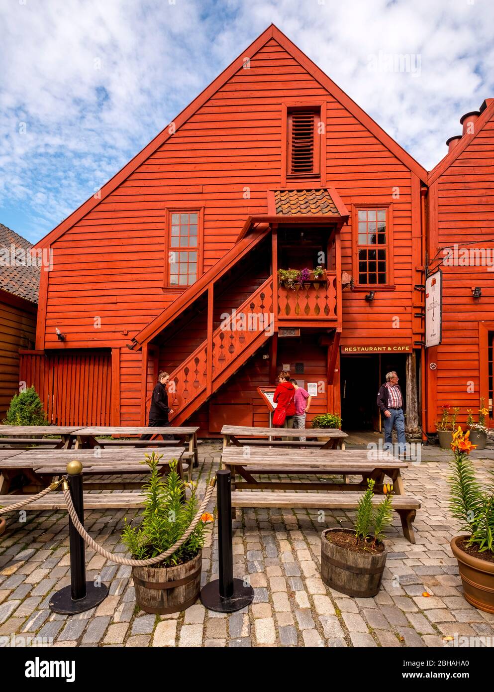 I turisti camminano di fronte ad una casa in legno scolpita con scala in legno, circondata da un cortile pavimentato sui tavoli e panche, decorato con vasche di fiori, a Bryggen, Bergen, Hordaland, Norvegia, Scandinavia, Europa Foto Stock