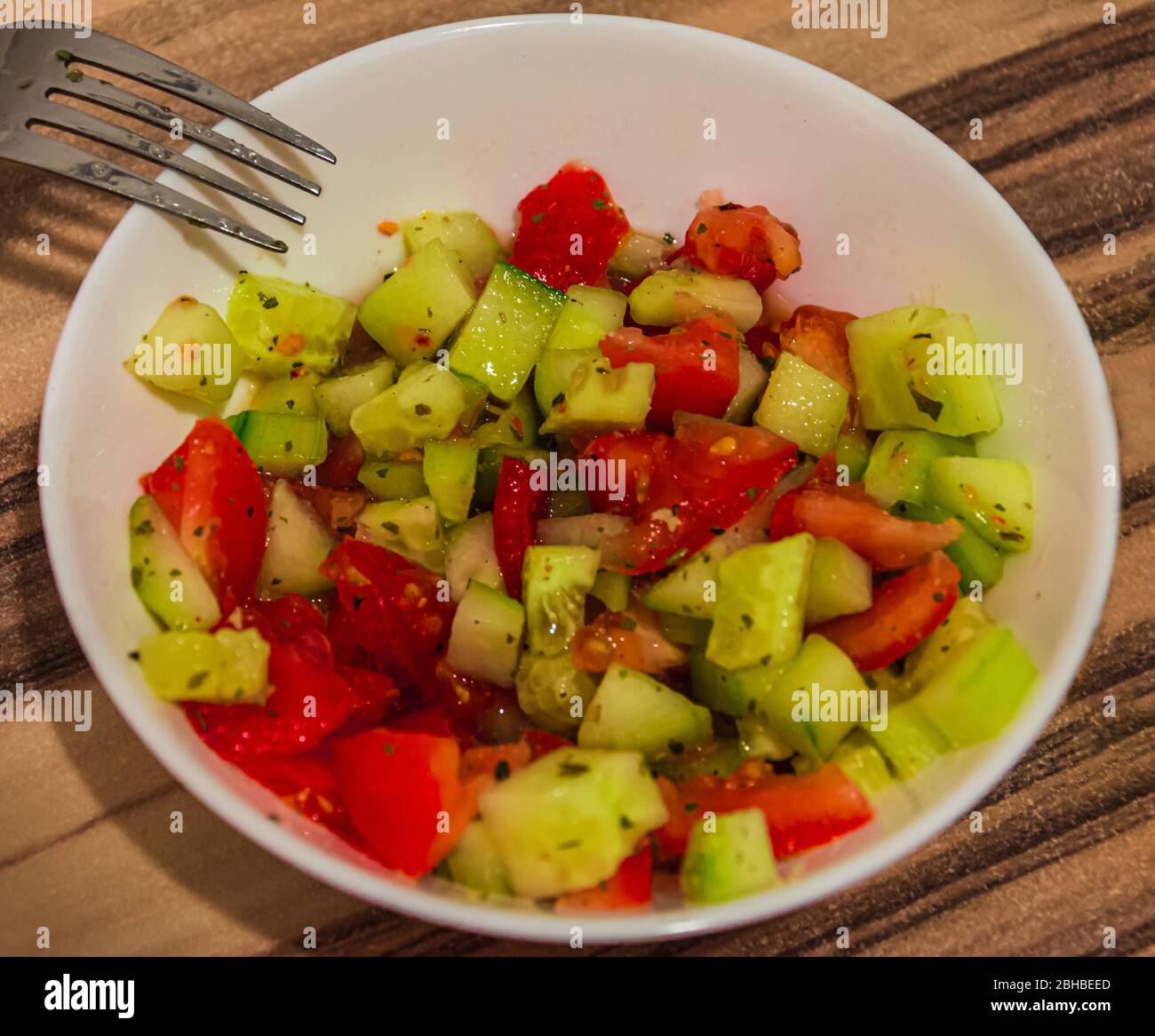 Insalata succosa di pomodori e cetrioli preparati con spezie in una ciotola bianca su un tavolo di legno Foto Stock